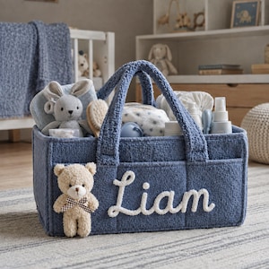 May include: A blue textured fabric storage basket with two handles, featuring the name Liam embroidered in white script on the front. A small beige teddy bear with a brown and white checkered bow is attached to the front, filled with baby items.