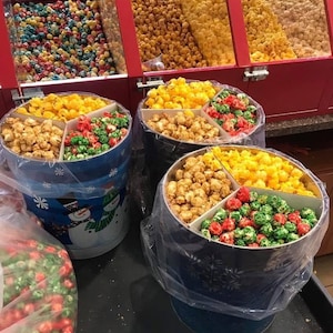 May include: A display of various popcorn flavors. Several bins hold popcorn in colors like yellow, caramel, red, green, and blue. Round containers with dividers showcase different popcorn varieties, including caramel and festive red and green.