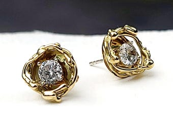 May include: A pair of gold stud earrings. Each earring features a round, clear gemstone set within a textured, organic-shaped gold setting. The earrings are displayed on a white surface.