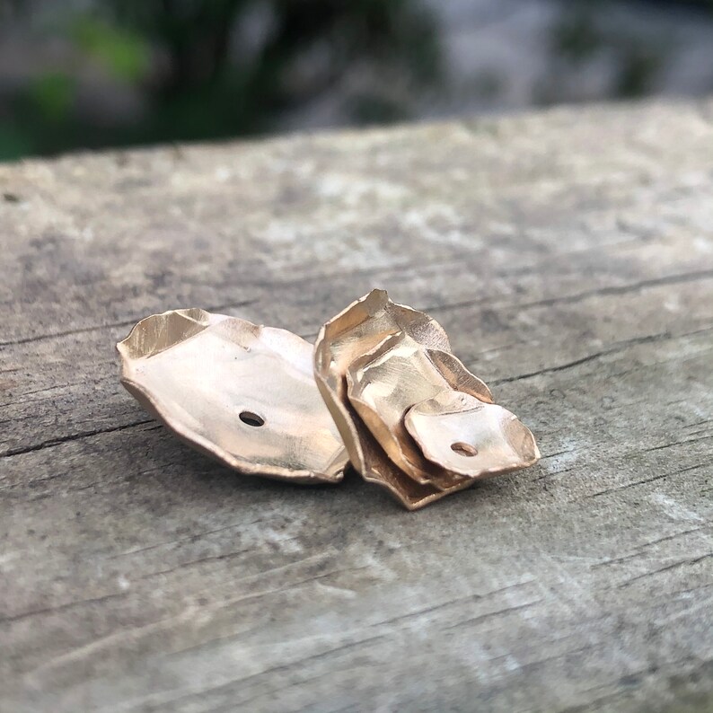 Może przedstawiać: Para złotych kolczyk&oacute;w o unikalnym, organicznym kształcie. Kolczyki wykonane są z wielu warstw metalu, co nadaje im fakturowany wygląd.