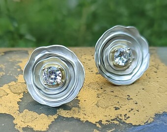May include: A pair of silver earrings with a round, clear gemstone set in a gold center. The earrings have a layered, textured design.