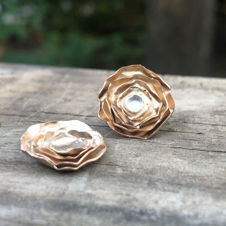 Może przedstawiać: Para złotych kolczyk&oacute;w z warstwowym, kwiatowym wzorem. Każdy kolczyk ma mały, przezroczysty kamień w środku.