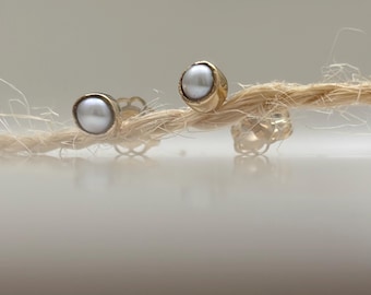 May include: A pair of stud earrings featuring round, white, lustrous pearls set in gold-toned metal bezels. The earrings include clear, scalloped-edge plastic earring backs, resting on a piece of light-colored, fibrous, twisted natural twine.