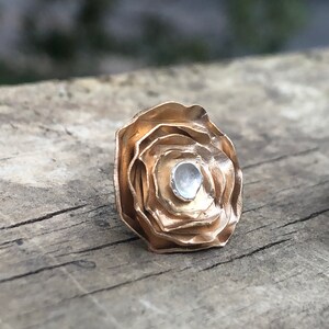 Może przedstawiać: Złota metalowa r&oacute;ża z srebrnym środkiem, płatki są warstwowe i fakturowane, r&oacute;ża leży na drewnianej powierzchni.