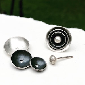 May include: A set of silver earrings with a black enamel finish. The earrings feature a circular design with concentric circles and a small sphere in the center. The earrings are shown disassembled, with the backings and posts separate from the fronts.