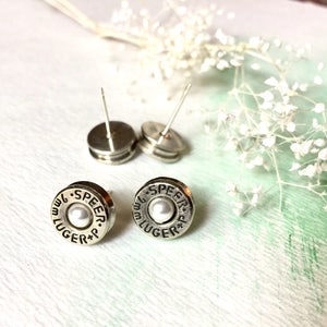 May include: A pair of silver bullet earrings with a white pearl in the center. The earrings are engraved with the text "9mm LUGER +P SPEER."