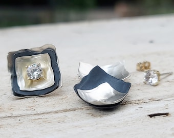 May include: A pair of layered square stud earrings featuring a central clear round gemstone, a silver-toned inner square, and a dark matte outer square. A separate pair of small clear round stud earrings with gold-toned posts is also visible.