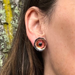 May include: A pair of round earrings with a layered design. The earrings are made of copper and have a black, orange, and red color scheme. The earrings have a small black stone in the center.