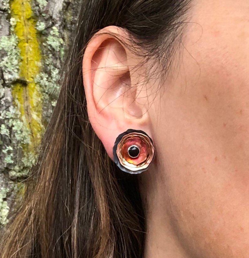 May include: A pair of round earrings with a layered design. The earrings are made of copper and have a black, orange, and red color scheme. The earrings have a small black stone in the center.