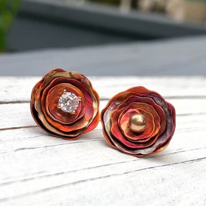 May include: A pair of copper earrings with a layered flower design. One earring features a small, clear gemstone in the center, while the other earring has a small, gold bead in the center.