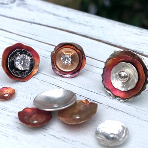 May include: A set of six copper and silver flower-shaped jewelry components with a clear rhinestone in the center of each. The components are arranged on a white wooden surface.