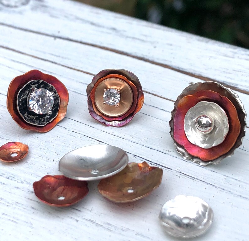 May include: A set of six copper and silver flower-shaped jewelry components with a clear rhinestone in the center of each. The components are arranged on a white wooden surface.