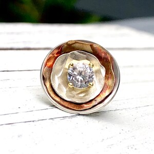 May include: A round, silver, copper, and gold ring with a clear gemstone in the center.