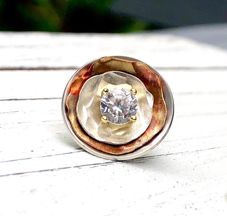 May include: A round, silver, copper, and gold ring with a clear gemstone in the center.