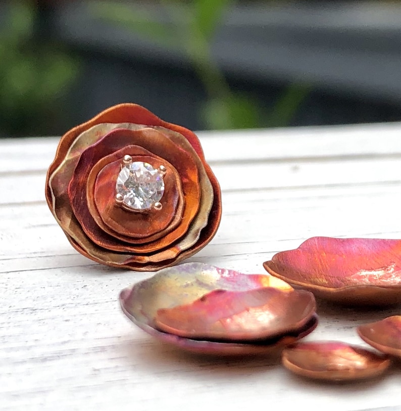 May include: A copper flower-shaped ring with a clear gemstone in the center. The ring is made of multiple layers of copper, creating a dimensional effect.