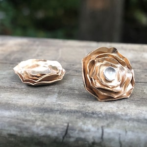 Może przedstawiać: Para złotych kolczyk&oacute;w w kształcie r&oacute;ż. Każdy kolczyk ma warstwowy wz&oacute;r z małym, okrągłym, srebrnym środkiem.