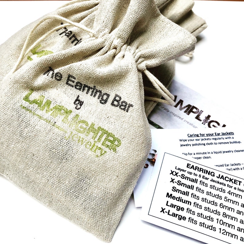 May include: A small, white canvas bag with a drawstring closure. The bag has the text "The Earring Bar by Lamplighter Jewelry" printed on it. A white card with the text "Caring for your Ear Jackets" and a size chart for earring jackets is visible in the background.