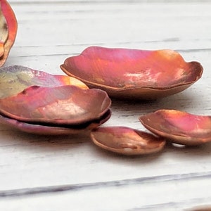 May include: A set of five copper petal-shaped dishes with a wavy edge. The dishes are iridescent with shades of pink, orange, and yellow.