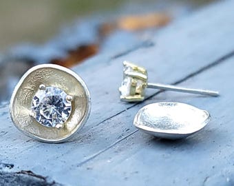 May include: Close-up of two silver stud earrings with a clear, round gemstone. One earring is in focus, showcasing the gemstone set in a textured, circular setting. The other earring is slightly out of focus, with a post and backing visible.