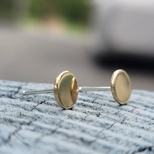May include: A pair of gold-toned stud earrings with a round, slightly domed shape. The earrings are on a wooden surface.