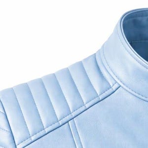 May include: Close-up of a light blue leather jacket. The jacket features a quilted shoulder design and a round collar. The stitching is visible, and the jacket appears to be made of a smooth material. The image is set against a white background.