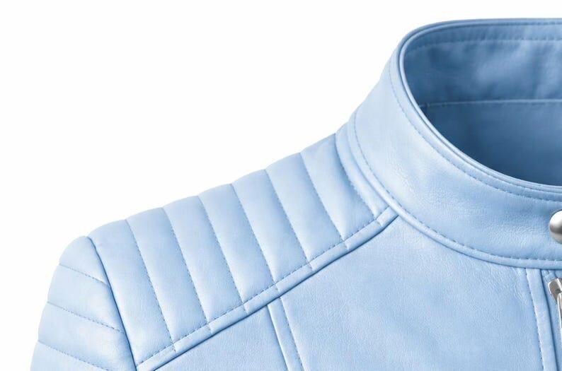 May include: Close-up of a light blue leather jacket. The jacket features a quilted shoulder design and a round collar. The stitching is visible, and the jacket appears to be made of a smooth material. The image is set against a white background.