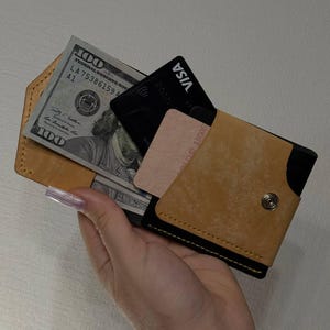 May include: A tan leather wallet with a black interior, holding a stack of $100 bills, a black Visa card, and a pink card. The wallet is secured with a snap closure.