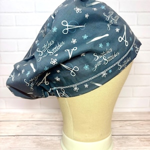 May include: A dark blue scrub cap featuring a repeating pattern of white scissors, tweezers, and small flowers. The fabric is printed with the text Snitches get Stitches in a white cursive font. The cap has a gathered design and a stitched hem.