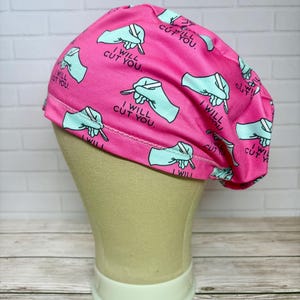 May include: A bright pink scrub cap featuring a repeating pattern of light blue gloved hands holding scalpels. The text "I WILL CUT YOU." is printed in black next to each graphic. The cap has a rounded crown and a stitched hem along the bottom edge.