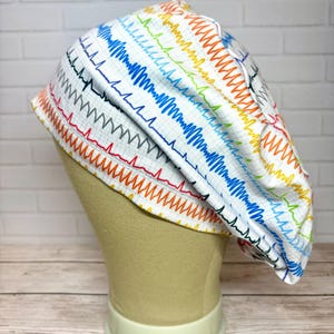 May include: A scrub cap featuring a white background with a grid pattern and a repeating print of colorful EKG heart rhythm lines in shades of blue, green, orange, and red. The fabric is gathered at the back with a rounded, bouffant-style shape.