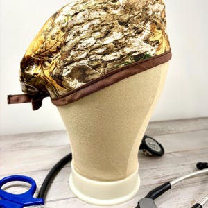May include: A scrub cap featuring a realistic forest camouflage pattern with brown, tan, and green leaf and bark motifs. The cap is finished with a solid dark brown fabric trim along the bottom edge and includes a tie closure at the back.