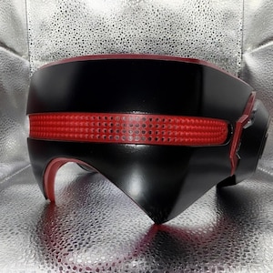 Puede incluir: Un casco negro con acabado mate liso que presenta una prominente banda roja horizontal en la parte frontal. La banda roja tiene una superficie texturizada y perforada. El casco tiene un diseño curvo y angular con detalles interiores rojos.