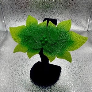 Pode incluir: Um acessório de cabelo com um grupo de folhas verde-vibrantes com uma superfície texturizada e nervurada. No centro do arranjo de folhas, há quatro pequenas contas verdes redondas e brilhantes agrupadas sobre uma base preta.