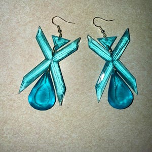 May include: A pair of turquoise earrings with a geometric design. Each earring features an X-shaped structure with a triangular top and a teardrop-shaped pendant. The earrings have gold-coloured fish hook ear wires.