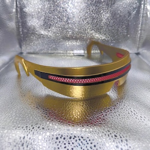May include: A gold-colored visor with a horizontal black band across the center. The band features a thin, perforated red strip running horizontally through its middle. The visor has a curved, wrap-around frame design with integrated side arms.