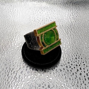 Puede incluir: Un anillo con una banda metálica oscura y texturizada y un marco rectangular dorado prominente. El centro del marco contiene un emblema circular verde con un patrón de líneas concéntricas, flanqueado por dos barras verdes a cada lado.