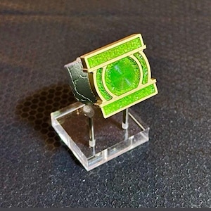 May include: A metallic ring featuring a rectangular gold-toned frame with a bright green textured inlay and a central circular green emblem with concentric circular detailing. The ring band has a dark, textured finish and is displayed on a stand.