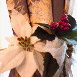May include: A decorative curtain tieback featuring a white poinsettia flower with red berries and green leaves. A wooden dowel holds the floral arrangement against patterned curtains. The Crafty Bug logo is in the upper left corner.