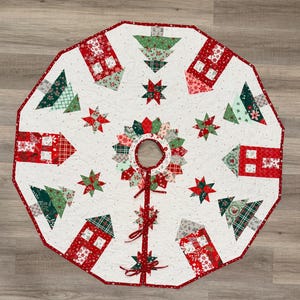 May include: A circular quilted Christmas tree skirt featuring a white background with red and green patchwork patterns. The design includes alternating house and tree motifs, star shapes, and a central opening with red fabric ties.