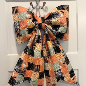 May include: A large fabric bow featuring a patchwork design of orange, black, and light green squares. The fabric is printed with Halloween-themed motifs including ghosts, bats, pumpkins, and stars. The bow has two long, pointed tails.