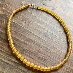 Könnte beinhalten: Eine Halskette aus kleinen, quadratischen, bernsteinfarbenen Perlen. Die Perlen sind aufgefädelt und bilden eine kreisförmige Form mit einem goldfarbenen Verschluss. Die Halskette wird auf einer Holzoberfläche präsentiert.