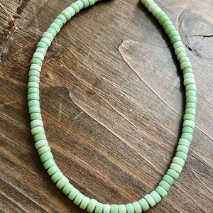 May include: A necklace featuring light green, disc-shaped beads. The beads are strung together with gold-colored wire and clasps. The necklace is displayed on a wooden surface.