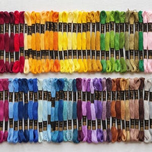May include: A collection of embroidery floss skeins arranged in two horizontal rows, featuring a full spectrum of colors including reds, oranges, yellows, greens, blues, purples, browns, grays, white, and black, each with a black paper label.