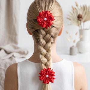 Op de afbeelding: Twee felrode stoffen bloemhaaraccessoires met gelaagde bloemblaadjes en een centraal cluster van kleine witte kraaltjes. Deze ronde bloemornamenten zijn bevestigd aan een blond gevlochten kapsel, geplaatst aan de boven- en onderkant van de vlecht.