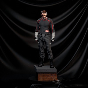 Puede incluir: Figura detallada de un hombre con una camisa gris oscura de manga larga con detalles rojos y pantalones a juego. La figura está sobre una base de ladrillo y hormigón. La figura tiene botas negras y manos vendadas.
