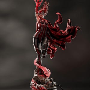 May include: A detailed statue of a female superhero in a dynamic pose, clad in a red and black costume with a flowing cape. She holds a red, circular object aloft, with her red hair flowing. The base features a glowing red energy effect.