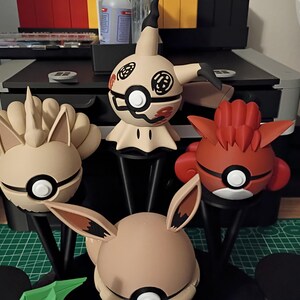 Op de afbeelding: Een verzameling Pok&eacute;mon-karakterbeeldjes in verschillende kleuren, waaronder beige, rood en wit. Elke figuur heeft een ronde basis met een zwarte standaard. De beeldjes worden tentoongesteld op een groene snijmat.