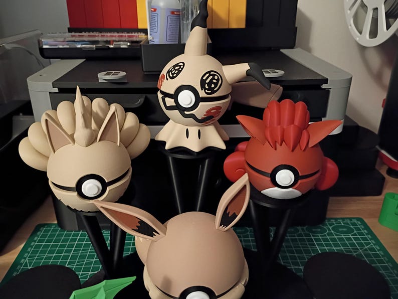 Op de afbeelding: Een verzameling Pok&eacute;mon-karakterbeeldjes in verschillende kleuren, waaronder beige, rood en wit. Elke figuur heeft een ronde basis met een zwarte standaard. De beeldjes worden tentoongesteld op een groene snijmat.