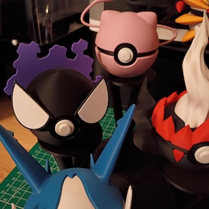 Op de afbeelding: Een verzameling kleurrijke, gestileerde figuren, waaronder een roze bol met katachtige oren, een zwart figuur met witte accenten en een rood-zwarte figuur met een witte, vlamachtige top. De figuren staan op zwarte standaards.