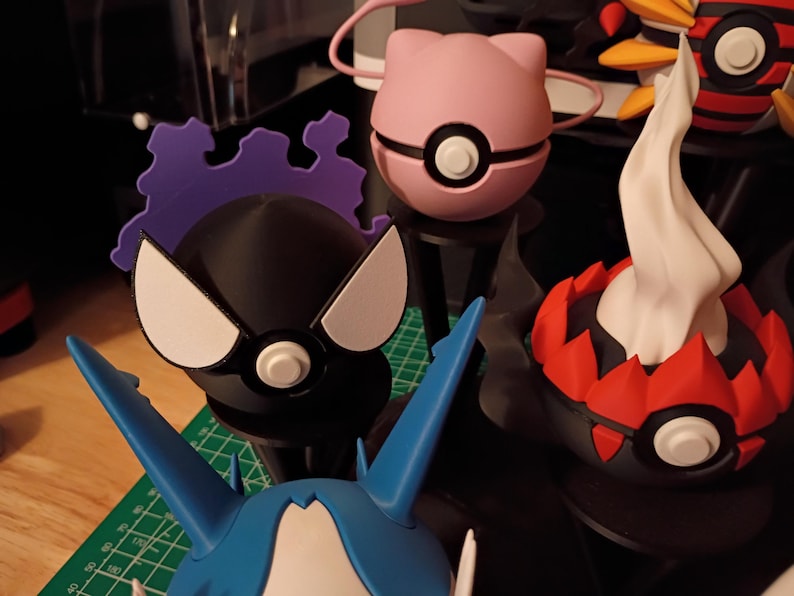 Op de afbeelding: Een verzameling kleurrijke, gestileerde figuren, waaronder een roze bol met katachtige oren, een zwart figuur met witte accenten en een rood-zwarte figuur met een witte, vlamachtige top. De figuren staan op zwarte standaards.